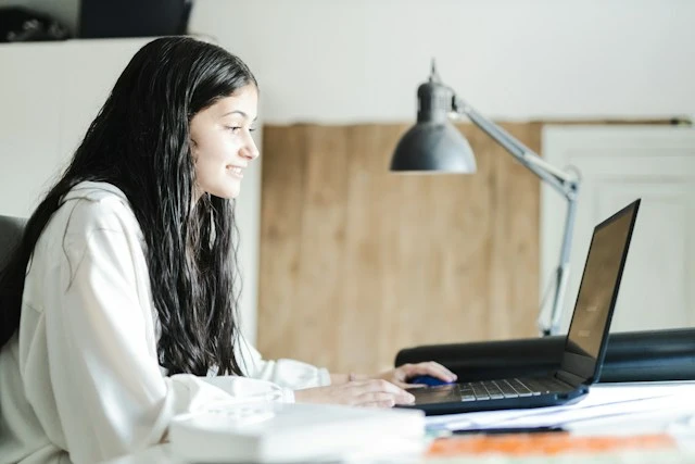 A young woman smiling as she works on her laptop at a desk.